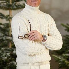 William Lockie, a man wearing a white William Lockie Ecru Cable Knit Pure Cashmere Rollneck sweater and glasses, poses in front of a Christmas tree adorned with twinkling lights. The luxurious cashmere threads of his William Lockie Ecru Cable Knit Pure Cashmere Rollneck sweater