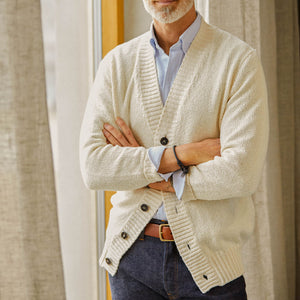 A man in an Ecru White Knitted Cotton Drumohr cardigan, blue striped shirt, and jeans standing with arms crossed.