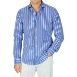 A man wearing a Fedeli dark blue striped cotton beach shirt with the sleeves rolled up, paired with white pants.