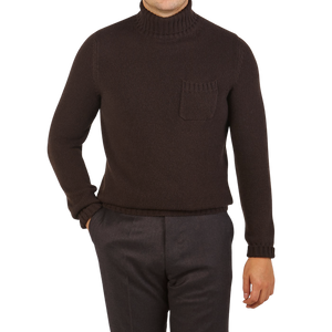 A person in a Fedeli Dark Brown Wool Cashmere Rollneck stands with one hand in their pocket wearing dark pants against a plain background.