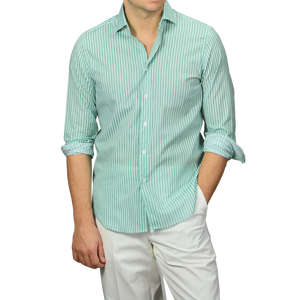 A person sporting a Fedeli Green White Striped Cotton Beach Shirt with rolled-up sleeves and slim-fit white pants stands with one hand in their pocket.