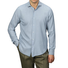 A person dressed in a Fedeli Light Blue Cotton Denim Shirt and olive green pants stands with one hand in their pocket against a plain gray background.