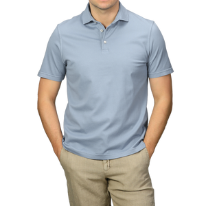 A person in a light blue organic cotton polo shirt by Fedeli and beige pants, showcasing luxury casual-wear, stands with hands in pockets against a gray background.