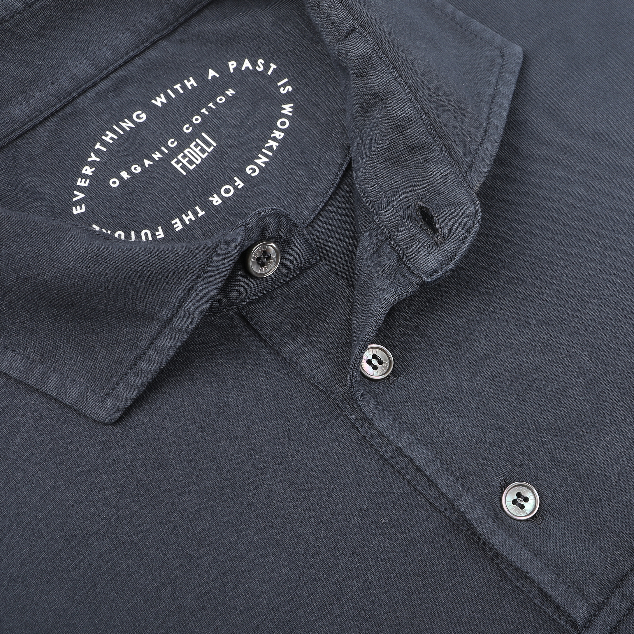 Close-up of a navy blue polo shirt collar made from organic cotton. The label inside reads: "Everything with a past is working for the future, organic cotton, FEEL," by Fedeli.