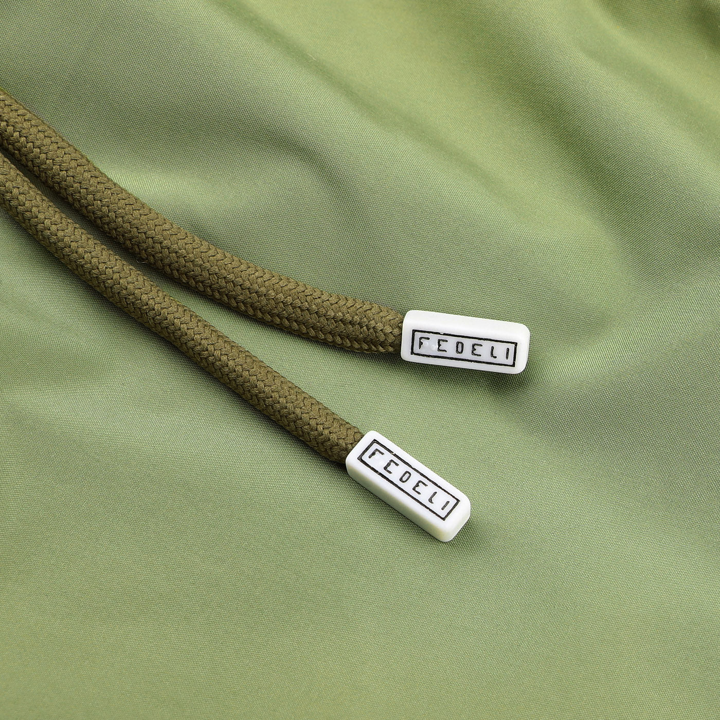 A close-up of two brown drawstrings with white tips labeled "Fedeli" on the Sage Green Madeira Microfiber Swim Shorts, made from quick-drying microfiber, highlights their luxurious and casual essence.