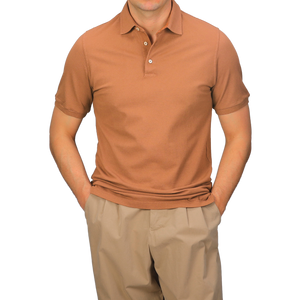 Wearing a Fedeli Washed Terracotta Cotton Pique Polo Shirt and beige pants, a person stands with hands in pockets against a gray background.