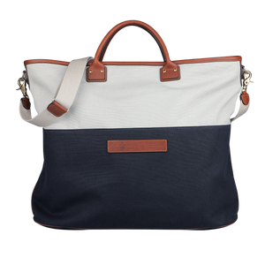 The Felisi Navy Grey Cotton Canvas Leather Travel Tote is crafted from durable cotton canvas, featuring brown vacchetta calf leather handles and a beige shoulder strap. It showcases a white upper section and a navy lower section, highlighted by a rectangular leather patch in the center.