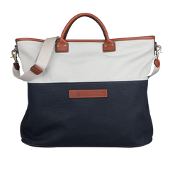 The Felisi Navy Grey Cotton Canvas Leather Travel Tote is crafted from durable cotton canvas, featuring brown vacchetta calf leather handles and a beige shoulder strap. It showcases a white upper section and a navy lower section, highlighted by a rectangular leather patch in the center.