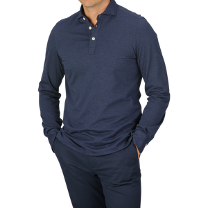 A person dressed in a Blue Melange Cotton Cashmere Jersey Polo Shirt by Finamore and dark pants, standing with hands in pockets. The polo shirt features a buttoned collar. The background is plain and light gray.