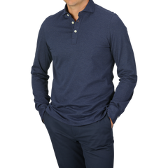 A person dressed in a Blue Melange Cotton Cashmere Jersey Polo Shirt by Finamore and dark pants, standing with hands in pockets. The polo shirt features a buttoned collar. The background is plain and light gray.