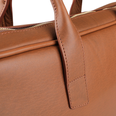 The image showcases the Frank Clegg Cognac Tumbled Leather Computer Briefcase, highlighting its fine stitching and a partially visible zipper. Crafted from tumbled calf leather, it offers a spacious interior designed to fit a 15" laptop.