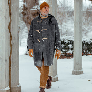A man in a Grey Melange Wool Monty Duffle Coat, made by Gloverall, walking in the snow.