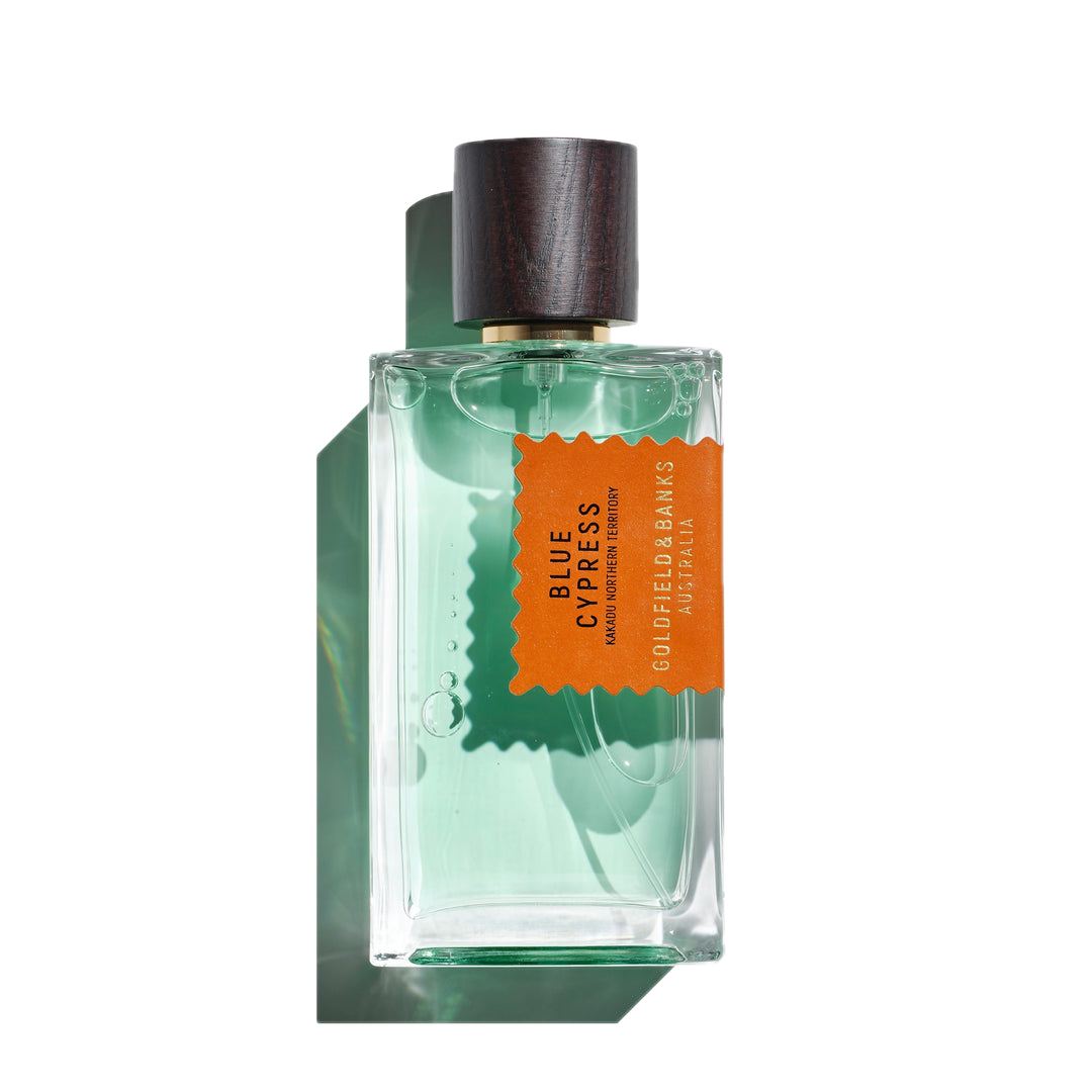 A 100ml Eau de Parfum glass bottle of Blue Cypress by Goldfield & Banks, showcasing a woody base with aromatic spices, topped with a wooden cap and containing a green-tinted liquid inside.