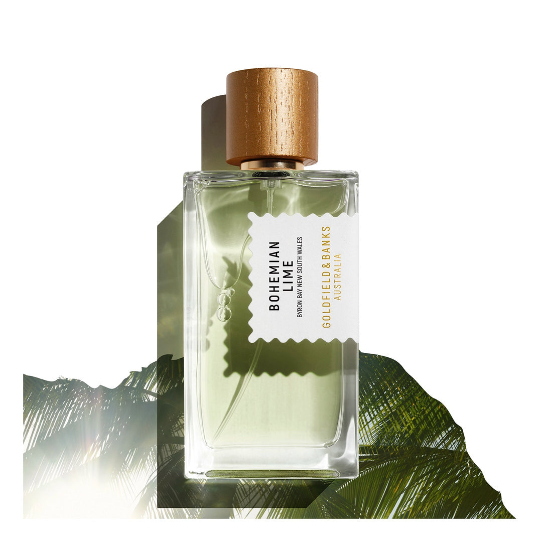 A glass bottle of Bohemian Lime 100ml Eau De Parfum from Goldfield & Banks, featuring a wooden cap, is gracefully set against a backdrop of green foliage, exuding fresh lime notes and earthy sandalwood.