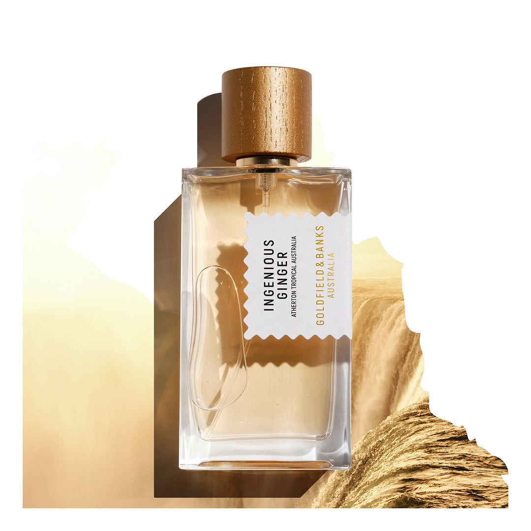 The Ingenious Ginger 100ml Eau De Parfum by Goldfield & Banks, featuring a clear glass bottle with a wooden cap, is showcased against a sandy, textured backdrop. The fragrance exudes notes of mandarin and warm amber.