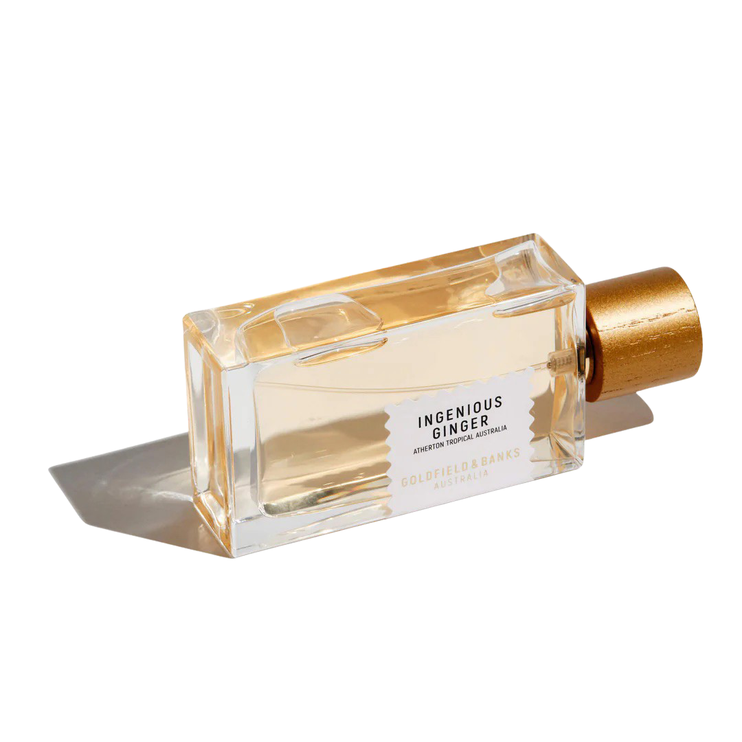 A clear rectangular perfume bottle of Goldfield & Banks' "Ingenious Ginger 100ml Eau De Parfum," adorned with a gold cap, rests on its side, casting a warm amber shadow.