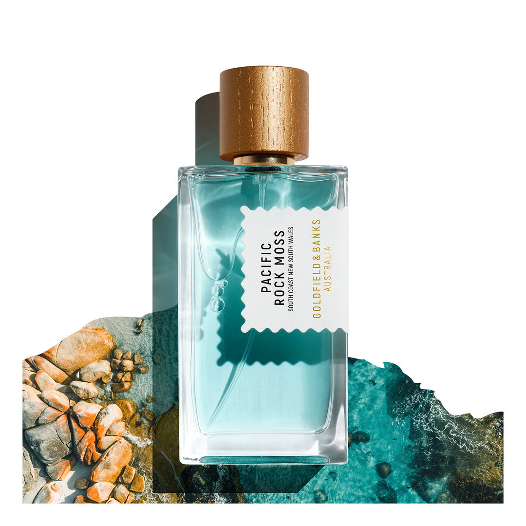 The "Pacific Rock Moss 100ml Eau De Parfum" by Goldfield & Banks, featuring a gold cap, encapsulates the essence of marine notes set against an ocean and rocky shoreline backdrop.