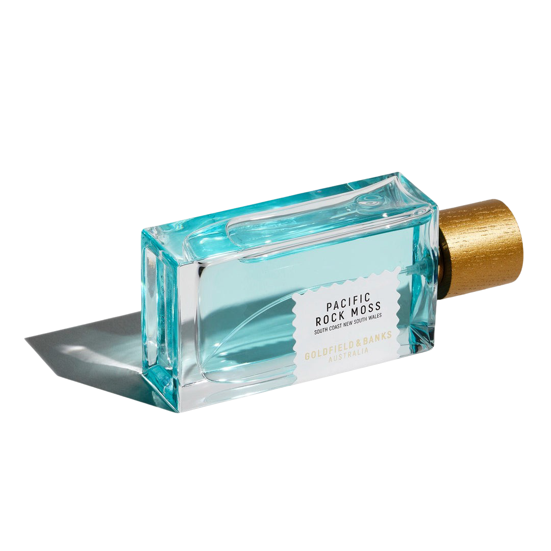 A rectangular bottle of Pacific Rock Moss 100ml Eau De Parfum by Goldfield & Banks, featuring a golden cap and filled with a light blue liquid that exudes a marine note, is lying on its side.