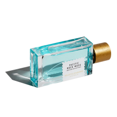 A rectangular bottle of Pacific Rock Moss 100ml Eau De Parfum by Goldfield & Banks, featuring a golden cap and filled with a light blue liquid that exudes a marine note, is lying on its side.