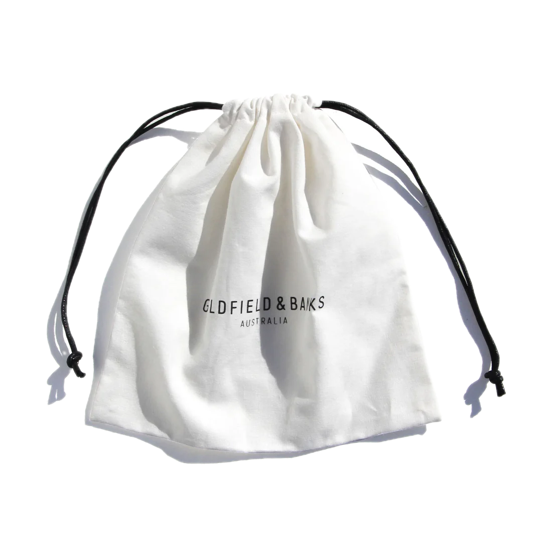 A white drawstring pouch featuring the "Goldfield & Banks" logo in black on the front conveys an essence akin to Blue Cypress, with hints of aromatic spices and a subtle woody undertone, perfectly encapsulating the 100ml Eau de Parfum by Goldfield & Banks.