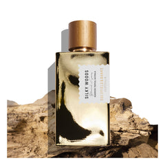 A gold perfume bottle of "Silky Woods 100ml Eau De Parfum" by Goldfield & Banks rests gracefully on a piece of driftwood against a white background, epitomizing the essence of luxury fragrance.