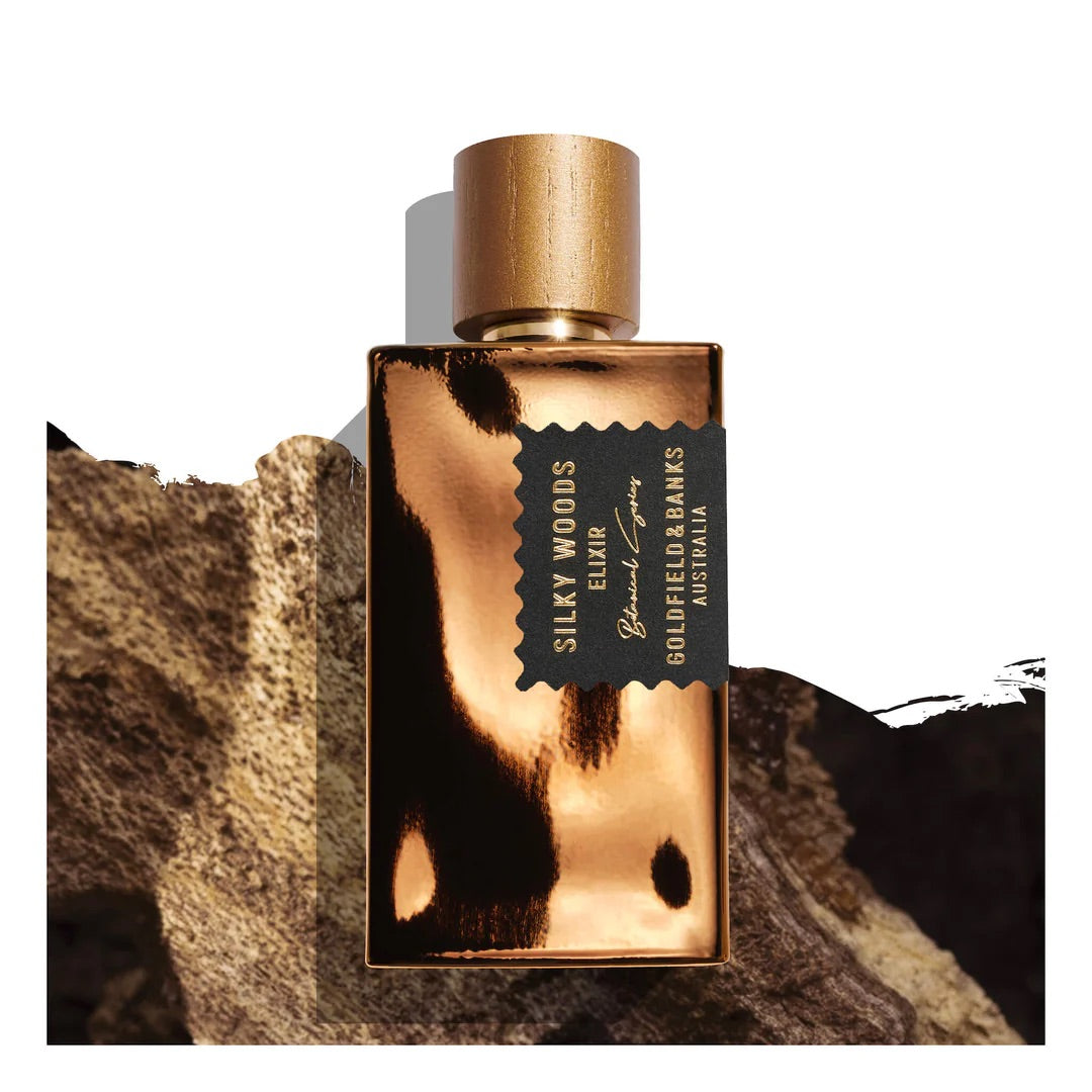 A rectangular gold bottle of "Silky Woods Elixir 100ml Extrait de Parfum" by Goldfield & Banks shimmers with an exotic botanical allure as it sits on a textured brown surface.