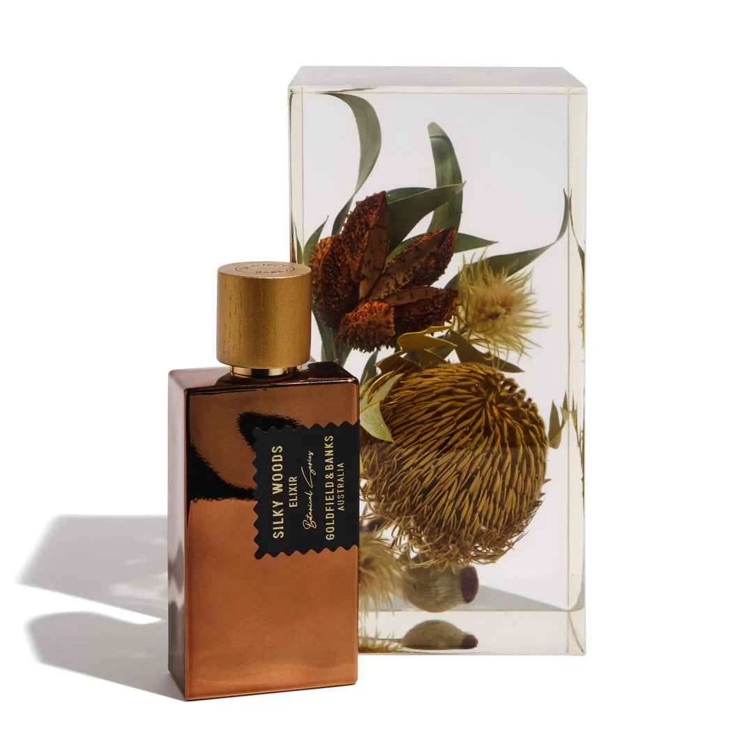 A bronze bottle of "Silky Woods Elixir 100ml Extrait de Parfum" by Goldfield & Banks is positioned next to a transparent block showcasing exotic dried botanical flowers.