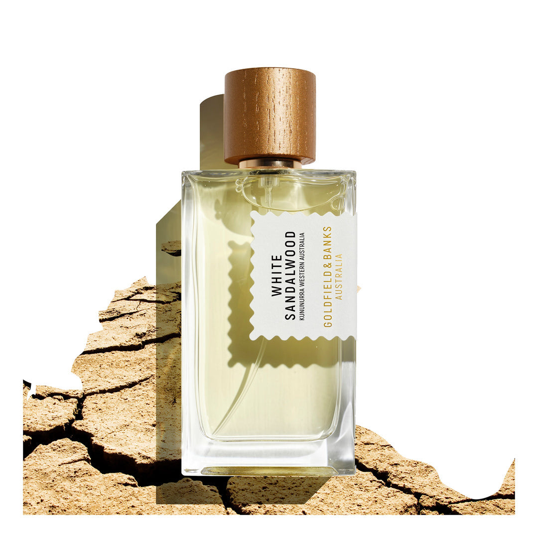 A 100ml bottle of Goldfield & Banks' White Sandalwood Eau De Parfum, featuring an elegant gold cap, is set against a cracked earth backdrop and exudes sophistication. Its scent is enriched with a subtle hint of amber, providing warmth and depth to the aromatic experience.