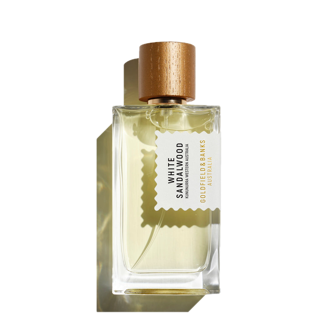 The White Sandalwood 100ml Eau De Parfum by Goldfield & Banks, presented in a clear glass bottle with hints of amber, is adorned with a gold cap and an elegant label, creating a delicate shadow.