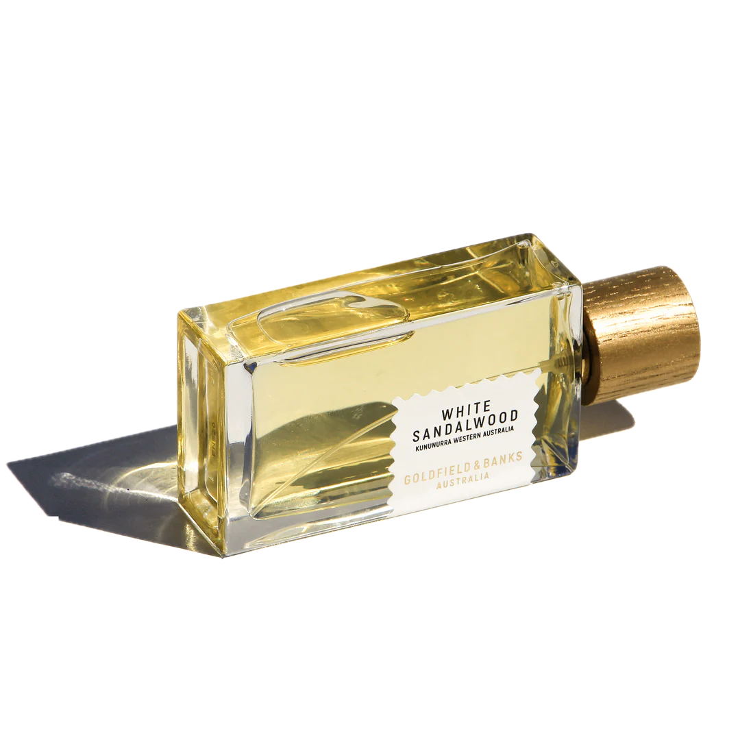 A 100ml bottle of Goldfield & Banks White Sandalwood Eau De Parfum, infused with hints of amber and adorned with a golden cap, rests elegantly on its side, casting a graceful shadow.