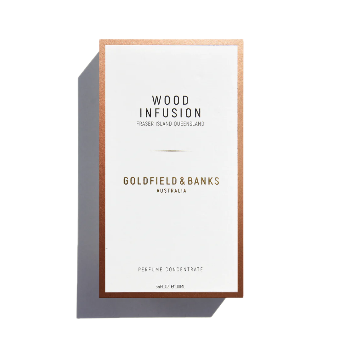 The packaging for "Wood Infusion 100ml Eau de Parfum" by Goldfield & Banks showcases the luxurious scent of Australian woods, including Sandalwood Album. The design features a white background with an elegant copper border.