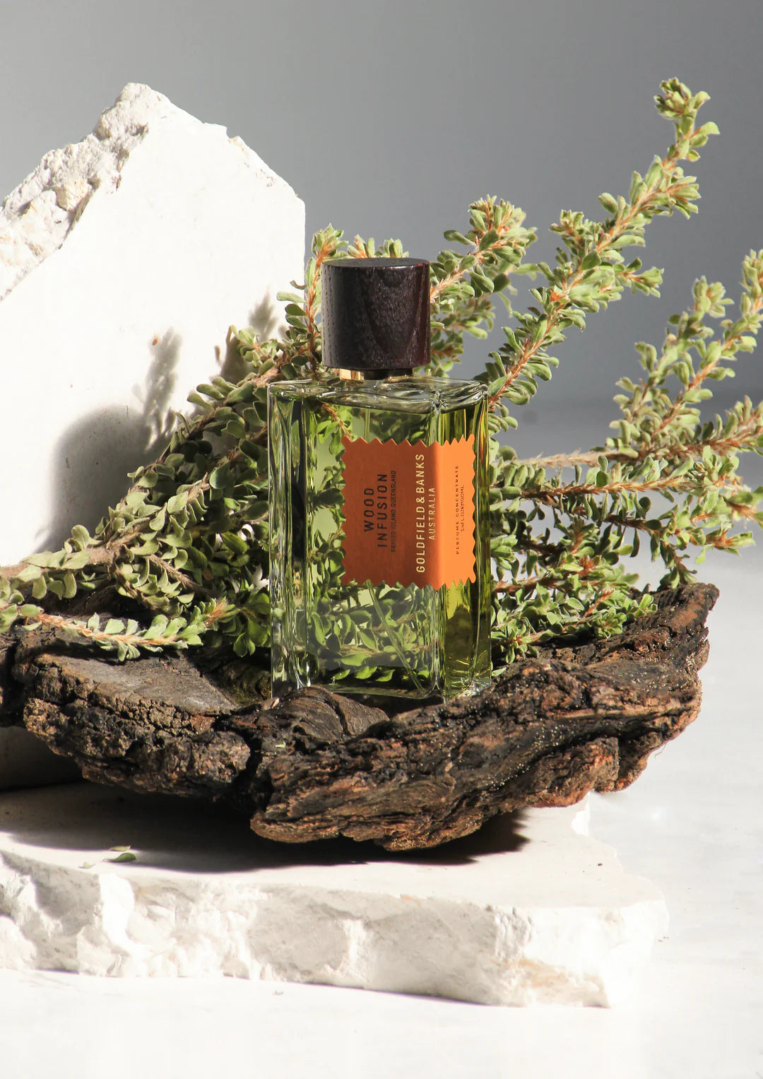 The "Wood Infusion" 100ml Eau de Parfum by Goldfield & Banks, presented in a glass bottle with an orange label, rests on a wooden surface amidst lush greenery and stone slabs, releasing the essence of Australian woods in its scent.