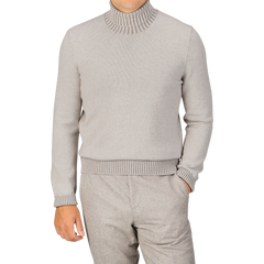 A person wearing a Beige Melange Pure Cashmere Rollneck sweater by Gran Sasso and matching light gray pants, with one hand in the pocket, standing against a plain background.