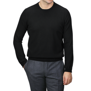 A person wearing the Black Cashmere Crewneck Sweater by Gran Sasso and gray pants stands with one hand in their pocket against a neutral background.