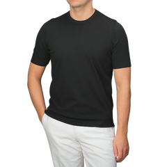 A man dressed in a slim-fit, Black Organic Cotton T-Shirt by Gran Sasso and white pants stands against a gray background.