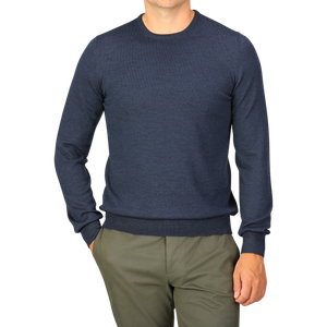 A person wearing a long-sleeve Blue Melange Extra Fine Merino Crew Neck by Gran Sasso with green pants stands with one hand in their pocket.