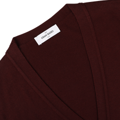 A close-up of the Bordeaux Extra Fine Merino Wool Cardigan reveals its exquisite detail: a maroon V-neck sweater with a white label that reads "Gran Sasso Italy.