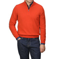 A person wearing a bright orange cashmere 1/4 zip sweater from Gran Sasso and dark blue jeans stands with one hand in a pocket, exuding the elegance of premium knitwear.