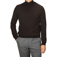 A person is wearing a Gran Sasso Brown Extra Fine Merino Roll Neck sweater and grey pants, with one hand in their pocket. They are facing the camera with squared shoulders, their face not visible.