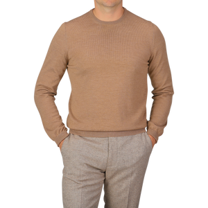 A person wearing a Camel Beige Extra Fine Merino Crew Neck by Gran Sasso and gray pants, with hands in pockets, standing against a plain backdrop.