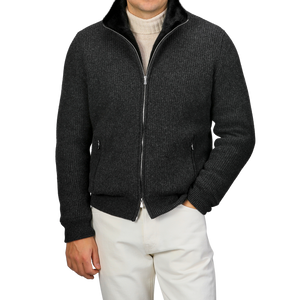 A person wearing a Charcoal Faux Fur Lined Cashmere Blouson by Gran Sasso over a light turtleneck sweater and white pants stands with hands in the jacket pockets against a plain background.