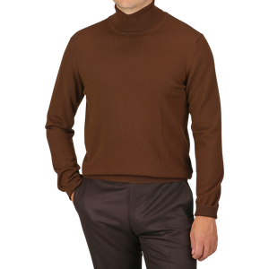 A person wearing a slim fit, Cinnamon Brown Extra Fine Merino Rollneck by Gran Sasso and dark brown pants stands with one hand in their pocket against a plain white background.