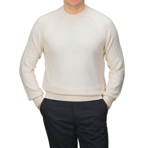 Dressed in Gran Sasso's Cream Cotton Boucle Crewneck Sweater and dark pants, a person with hands in pockets stands against a plain grey background. The pure cotton boucle fabric of the sweater adds texture to the simple outfit.