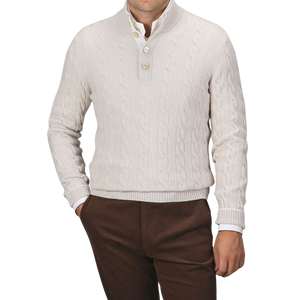 A man is wearing a cream Pure Cashmere Quarter Button Sweater by Gran Sasso over a white button-up shirt, paired with brown trousers. The top button of the cable-knitted sweater is undone. The background is plain gray.