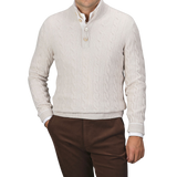 A man is wearing a cream Pure Cashmere Quarter Button Sweater by Gran Sasso over a white button-up shirt, paired with brown trousers. The top button of the cable-knitted sweater is undone. The background is plain gray.