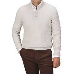 A man is wearing a cream Pure Cashmere Quarter Button Sweater by Gran Sasso over a white button-up shirt, paired with brown trousers. The top button of the cable-knitted sweater is undone. The background is plain gray.