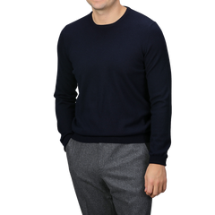 A person wearing a Dark Navy Cashmere Crewneck Sweater by Gran Sasso, along with gray trousers, stands against a plain background with hands in pockets.