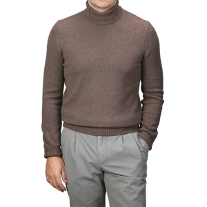 A person dressed in a Gran Sasso Fawn Brown Air Wool Rollneck and gray pants stands with one hand in a pocket against a plain backdrop.