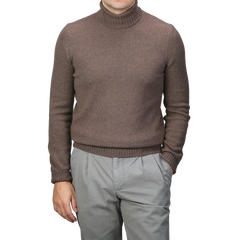 A person dressed in a Gran Sasso Fawn Brown Air Wool Rollneck and gray pants stands with one hand in a pocket against a plain backdrop.