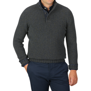 A person wearing a Gran Sasso Green Melange Wool Quarter Button Sweater paired with dark blue trousers is standing with one hand in a trouser pocket against a grey background.